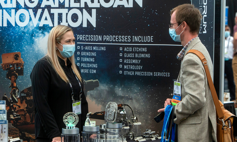 Two people having a conversation wearing masks in front of a company backdrop