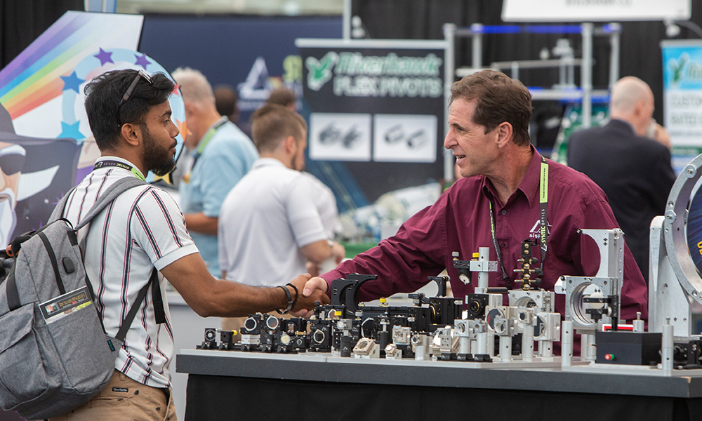 Exhibitor shaking hands with an attendee in the exhibit hall reaching across a table full of products
