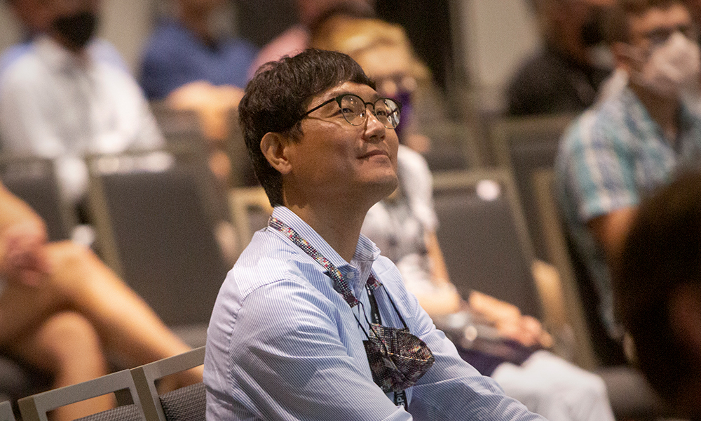 In a crowd, a male with glasses tilts head and smiles, while listening to an EDI presenter