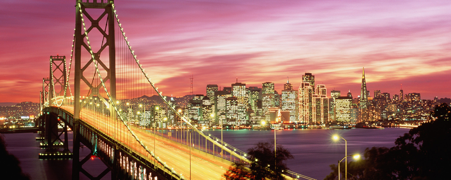 The Bay Bridge in the foreground with San Francisco city lights and a colorful California sunset in the background