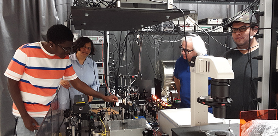 Experimental setup for a magnetometer using alkali sodium atoms in Professor Tripathi's Delaware State University lab. The yellow glow from a sodium laser is visible in the picture. Left to right: Lawrence Taylor, a freshman pursuing a BS in Engineering Physics; Tripathi; Robin Depto, a BS Electrical Engineering student at Delaware Technical Community College; and Mauricio Pulido who pursuing an MS in Applied Optics.